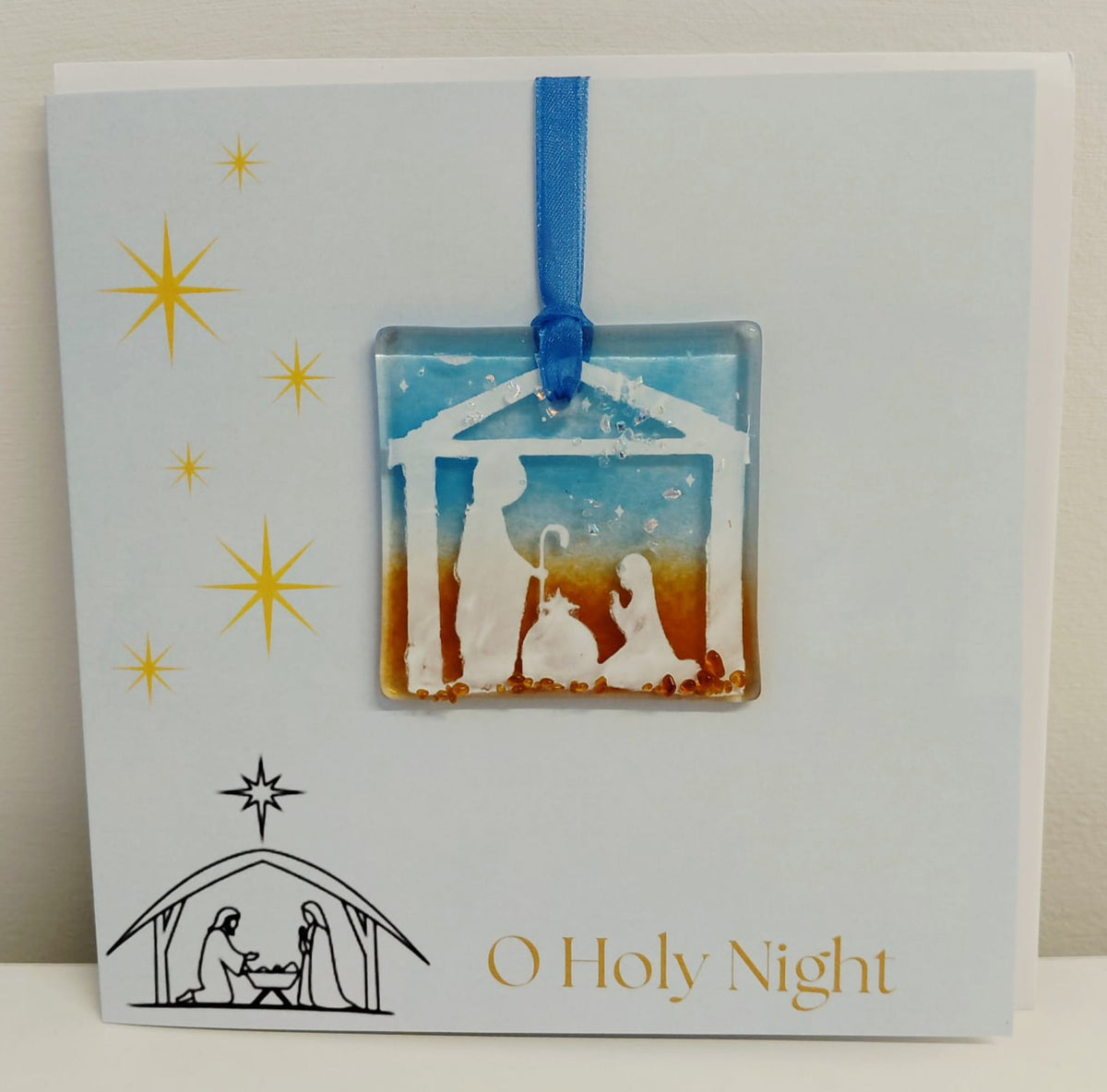 Handcrafted 6x6 inch “O Holy Night” Christmas greeting card featuring a detachable square fused glass nativity ornament with Joseph, Mary, baby Jesus in a manger silhouette against a sky blue and warm amber background.