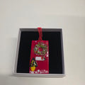 A small red Christmas ornament with a green wreath, white candle, and black-and-yellow ornament, displayed inside a black-lined gift box on a white wall.
