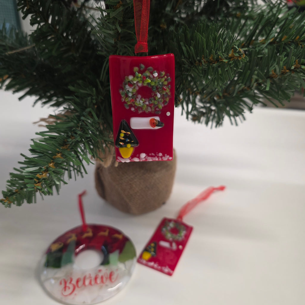 A red rectangular Christmas ornament hanging on a green artificial tree, featuring a wreath, a candle, and a small bird. Another red ornament tag and a round clear ornament are visible nearby against a white background.